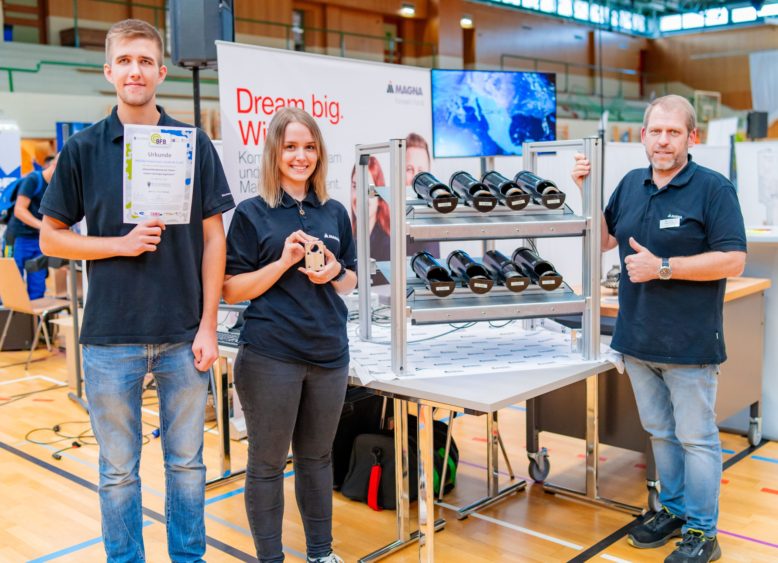 Zwei Lehrlinge vor dem Messestand von Magna mit der Urkunde in der Hand. Rechts daneben der Messeaussteller.